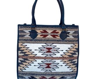 May include: A rectangular tote bag with a Southwestern-inspired pattern. The bag features a woven design in shades of white, beige, brown, and navy blue. It has navy blue leather handles and trim, and metal hardware.