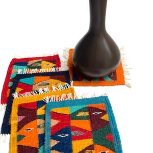 Puede incluir: Un jarrón de cerámica marrón oscuro se encuentra sobre un posavasos tejido colorido. Varios posavasos más con diseños geométricos de pájaros en rojo, naranja, amarillo y azul están dispuestos alrededor del jarrón. Los posavasos tienen flecos blancos.