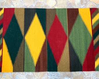 May include: A colorful woven rug with a geometric diamond pattern in shades of red, yellow, green, and brown. The rug has fringe on the ends.