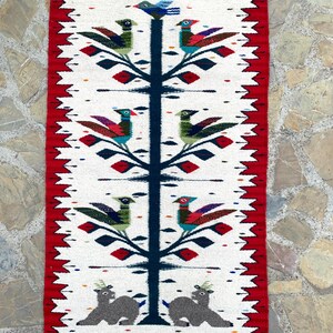 May include: Handwoven rectangular rug with a white background and a red border. The design features a central tree with colorful birds and deer at the base. The birds have blue, green, red, and purple details. The rug has a fringed edge.