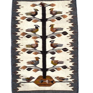 May include: Handwoven rectangular rug featuring a tree design with birds. The rug has a cream-colored background with a dark gray border and fringe. The tree is brown, and the birds are in shades of brown, gray, and yellow.