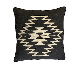 May include: Square black throw pillow with a geometric cream-colored design. The pattern features a series of zigzags and diamond shapes, creating a symmetrical design. The pillow is made of a woven fabric.