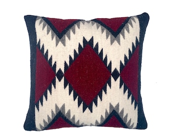 May include: A decorative pillow with a geometric pattern in shades of burgundy, black, white, and gray. The pillow has a diamond shape in the center and a border around the edges.