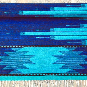 May include: A blue and turquoise woven rug with a geometric pattern. The rug has a fringe on the bottom edge.