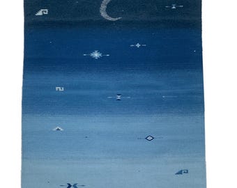 May include: A rectangular, handwoven rug with a gradient of blue hues, transitioning from dark indigo at the top to a light sky blue at the bottom. The rug features a crescent moon and scattered star-like patterns. Tassels are visible at the corners.