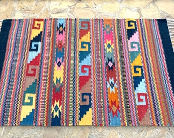 May include: A colorful woven rug with geometric patterns in shades of red, blue, yellow, and green. The rug has a black border and white fringe.
