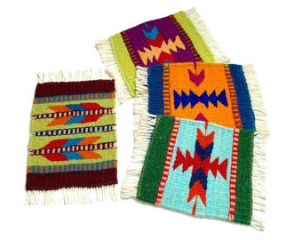 May include: Four colorful woven coasters with fringed edges. The coasters feature geometric patterns in shades of green, red, orange, blue, and purple. Each coaster is approximately 4 inches square.