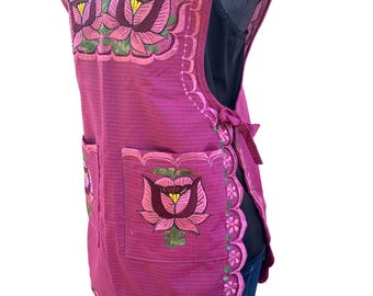May include: A magenta apron with embroidered floral designs. The apron features two pockets and a scalloped edge. The floral embroidery includes pink, green, and yellow accents. The apron is tied at the sides.