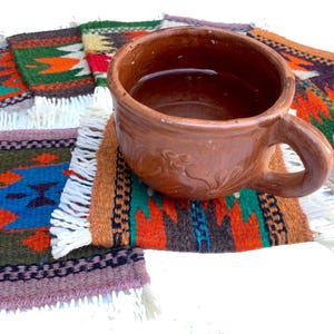 May include: A terracotta mug with a handle sits on a colorful woven rug with fringe. The mug is filled with a brown liquid.