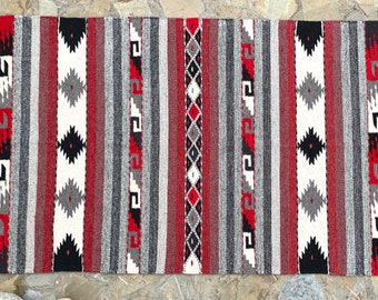May include: A red, white, black, and gray striped rug with geometric patterns. The rug has fringe on the ends.