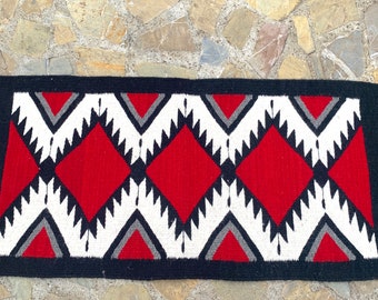 May include: Rectangular woven rug with a black border and fringe. The rug features a repeating pattern of red diamonds, white zigzags, and grey triangles. The rug is displayed on a stone surface.