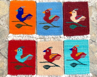 May include: Six colorful woven coasters featuring a stylized bird design. Each coaster is a different color and features a bird with a different color scheme. The coasters are made of a soft, textured material and have a fringe border.