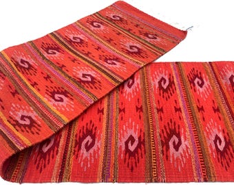 May include: A vibrant, handwoven runner in shades of red, orange, and brown. The textile features a repeating pattern of stylized motifs and stripes. The runner is made of a thick, textured material, perfect for adding warmth and color to any space.