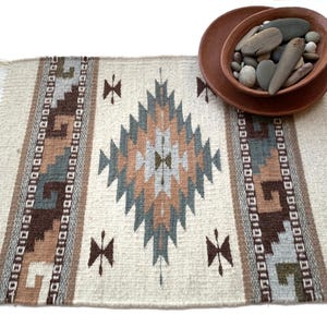 Puede incluir: Una alfombra tejida con fondo crema y un patrón geométrico en tonos marrón, azul y beige. La alfombra tiene un borde con flecos y se combina con un cuenco de terracota lleno de piedras.