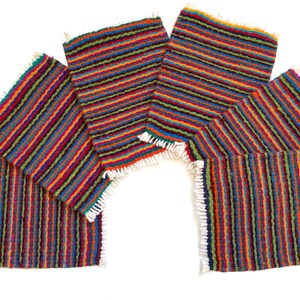 Puede incluir: Un conjunto de manteles individuales tejidos a rayas de colores. Los manteles individuales presentan rayas verticales en tonos rojos, naranjas, verdes y azules. Cada mantel individual tiene un borde con flecos. La disposición sugiere una decoración de mesa.