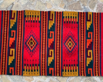 May include: A rectangular, handwoven rug with a vibrant red central field. The rug features geometric patterns in orange, yellow, and dark blue, with diamond-shaped motifs. White fringe adorns the short ends, adding a textural contrast. This rug is suitable for home decor.