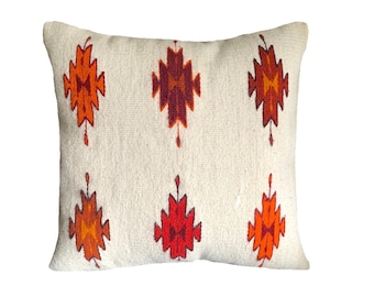 Puede incluir: Cojín decorativo crema con un patrón geométrico en tonos naranja y rojo. El cojín presenta un diseño repetitivo de formas de diamantes estilizadas, agregando un toque del suroeste a la decoración del hogar.