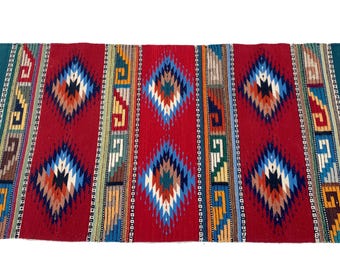 May include: A rectangular woven rug with a red background and geometric diamond patterns in blue, white, orange, and black. The rug has teal green borders and white fringe on the ends. A decorative textile for home decor.