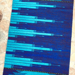 May include: A blue and turquoise woven rug with a geometric pattern. The rug has a fringe border.