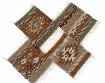 May include: A set of four square woven coasters featuring geometric patterns in shades of brown, tan, olive green, and cream. Each coaster has a textured surface and white fringe along two edges, arranged in a cross-like formation on a white background.