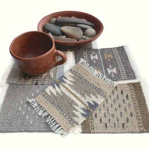 May include: A set of rectangular woven coasters featuring various geometric patterns in shades of gray, beige, and brown, each finished with white fringe on two sides. A brown clay mug and a brown clay bowl filled with smooth gray stones sit atop.
