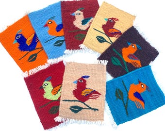 May include: Eight colorful woven coasters featuring stylized birds on branches. The coasters are made of wool and have a fringed edge.