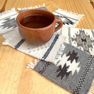 May include: Four woven coasters with fringe edges and geometric patterns in shades of gray and white. A brown ceramic mug with a dark brown liquid sits on top of the coasters.