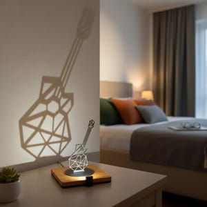 Peut inclure: Lampe en forme de guitare géométrique en métal blanc, projetant une ombre sur le mur. La lampe est posée sur un socle en bois, créant un effet lumineux unique. L'ombre de la guitare est projetée sur le mur. La lampe est sur une table de chevet.