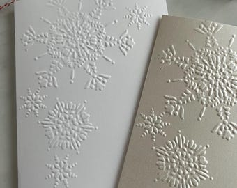 May include: Two folded greeting cards with embossed snowflake designs. One card is white, the other is a light beige. The snowflakes are detailed and textured, with various sizes. The cards are likely for winter holidays.