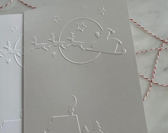 May include: Two embossed Christmas cards with a Santa Claus and reindeer design. The cards are light gray and white, with a textured surface. The design includes stars, a moon, houses, and trees. Red and white twine and silver bells are in the background.
