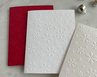 May include: Three embossed snowflake design greeting cards in red and white. The cards are rectangular and feature a raised snowflake pattern. Decorative bells and red and white twine are also visible.