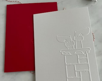 May include: Two rectangular Christmas cards. One is bright red, the other is white with an embossed chimney design. Red and white twine and silver bells are also visible, suggesting a holiday theme.