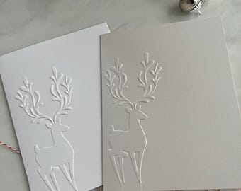 May include: Two blank greeting cards with embossed reindeer designs. One card is white, the other is light gray. Silver and gold jingle bells are scattered nearby, along with red and white twine. A festive holiday theme.