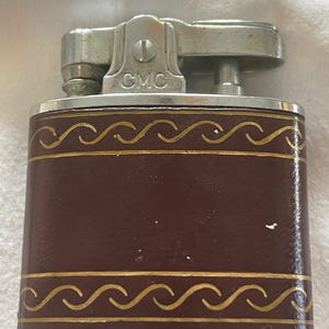 May include: A rectangular metal lighter with a dark brown finish and gold-colored wave patterns along the top and bottom edges. The silver-toned top mechanism features the letters GMC embossed on the front center of the metal casing.