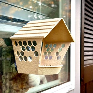 May include: A tan-colored bird feeder with a house-like design, featuring a sloped roof and honeycomb-shaped cutouts. The feeder is mounted on a window, offering a view of the outside. The feeder has a small tray for birdseed.