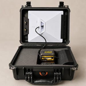 May include: A black hard-shell protective carrying case with a hinged lid, containing a white rectangular electronic device mounted inside the lid and a yellow and black DeWalt battery pack connected by a black cable in the foam-lined base.