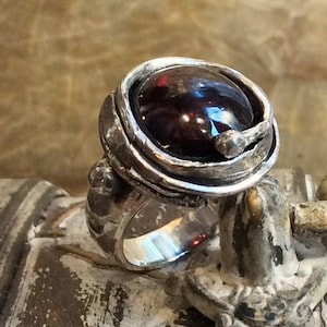 May include: A silver ring with a dark red gemstone set in a textured silver bezel. The ring has a unique, organic design with a twisted band.