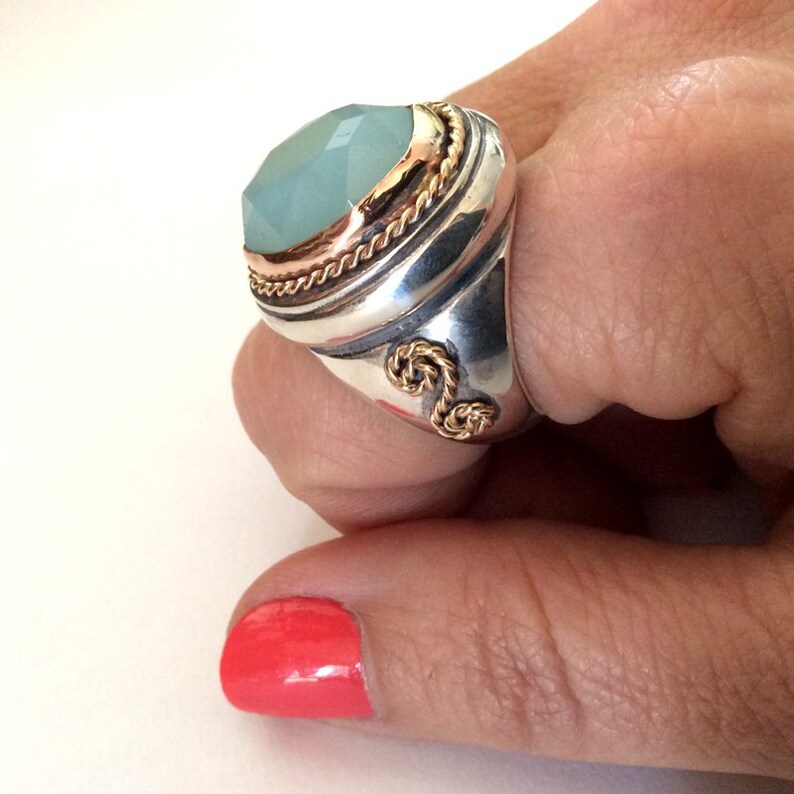 May include: A silver ring with a large, light blue gemstone set in a gold bezel. The ring has a rope-like design around the bezel and a twisted rope design on the band.