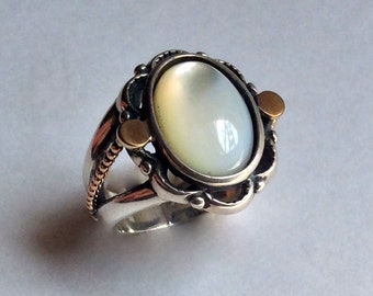 May include: A silver ring with a white oval gemstone set in a silver and gold filigree design. The ring has two gold accents on the sides.
