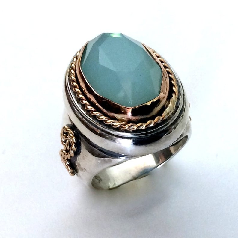May include: A silver ring with a large, oval-shaped, light blue gemstone set in a gold bezel. The ring has a rope-like design around the bezel and a decorative detail on the side.