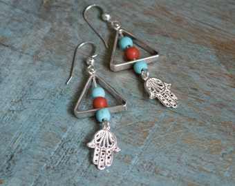 May include: Silver earrings with a triangle design and turquoise and red beads. The earrings have a hamsa charm hanging from the bottom.
