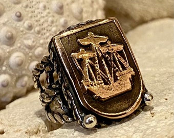 May include: A silver ring with a rope-like band and a square-shaped bezel featuring a gold-toned ship design.