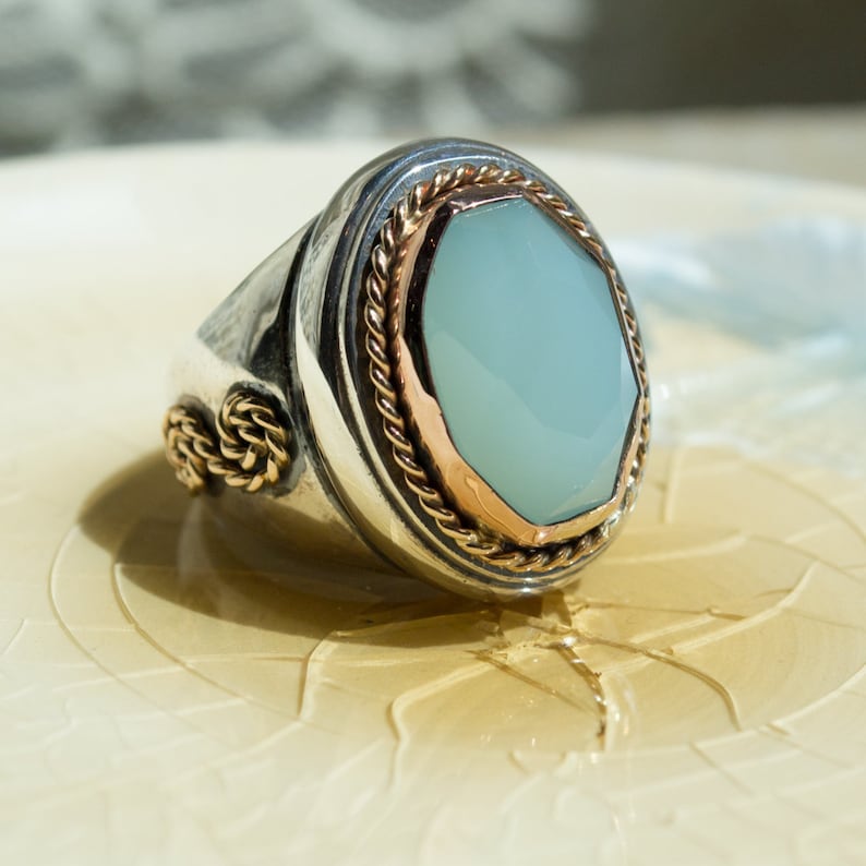 May include: A silver ring with a light blue gemstone set in a gold bezel. The ring is decorated with a rope-like design around the bezel.