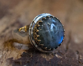 May include: A silver ring with a large, round, gray gemstone set in a gold filigree bezel. The ring is on a wooden surface.