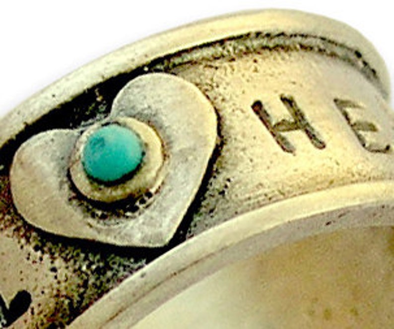 May include: Silver ring with a turquoise stone set in a heart shape. The ring is engraved with the word "HOPE".