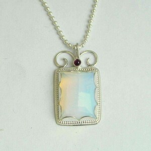 May include: A silver necklace with a rectangular pendant featuring a white opal gemstone and a small red garnet accent. The pendant has a delicate, ornate design.