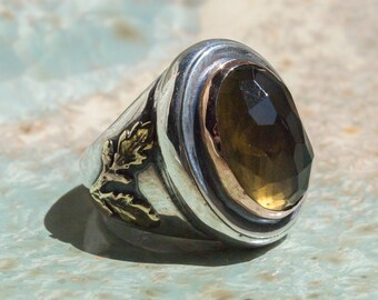May include: A silver and gold ring with a large, oval-shaped, faceted, brown gemstone. The ring has a decorative design of gold leaves on the side.