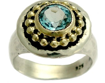 May include: A silver ring with a blue gemstone set in a gold bezel. The ring has a textured band and a decorative border of gold beads.