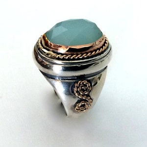 May include: A silver ring with a large, oval-shaped, light blue gemstone set in a gold bezel. The ring has a rope-like design around the bezel and two gold knots on the band.