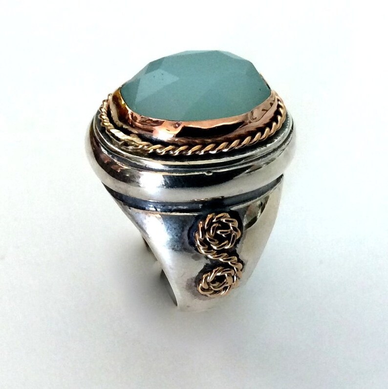 May include: A silver ring with a large, oval-shaped, light blue gemstone set in a gold bezel. The ring has a rope-like design around the bezel and two gold knots on the band.
